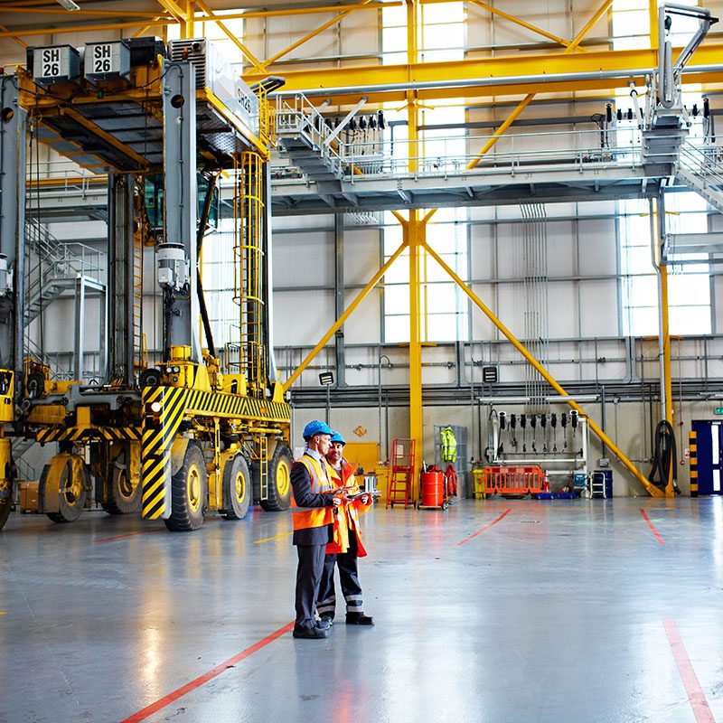 Two workers wearing blue hardhats, safety vests and reflective gear chat in a large industrial building