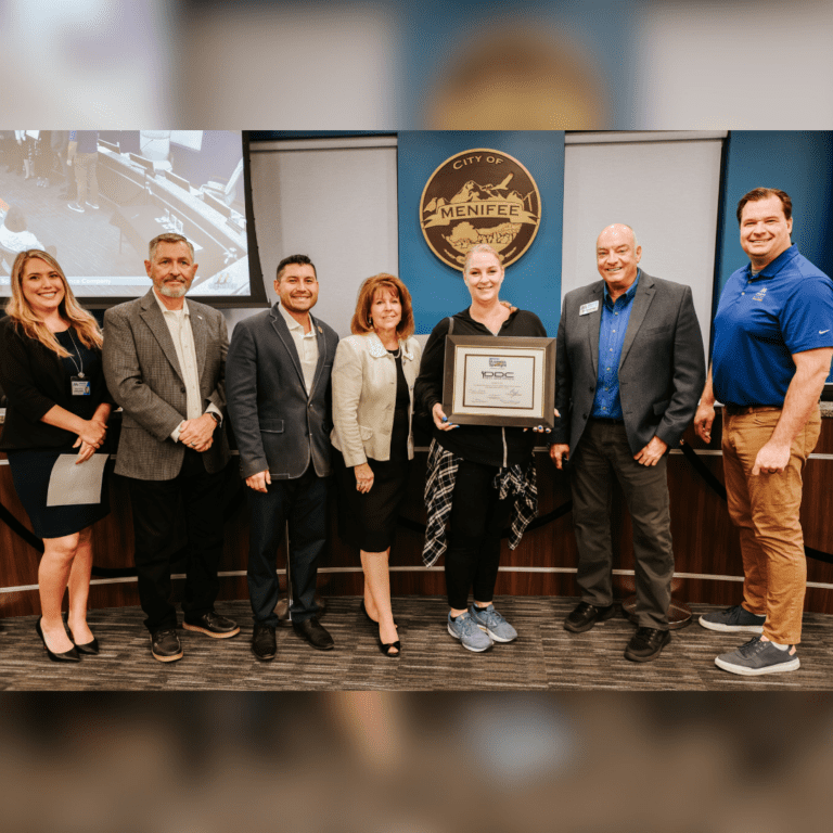 A group of seven people standing indoors, with one person in the center holding a framed certificate. A "City of Menifee" logo is visible in the background, highlighting Diana’s Dance Company as the December Business Spotlight.