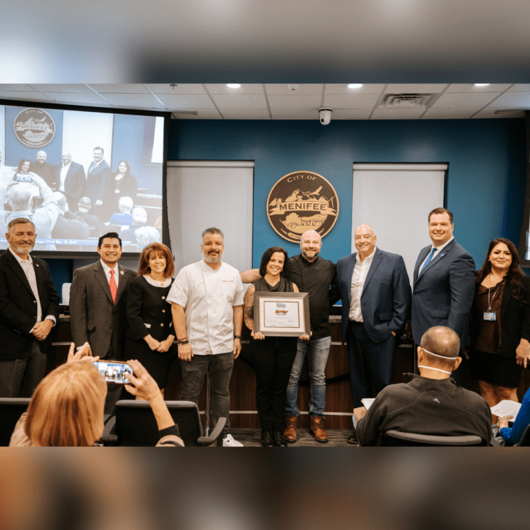 A group of people stands in a conference room, with one person holding a framed certificate. The background features a "City of Menifee" emblem on the wall. Audience members are seated, one taking a photo, capturing the moment as Savage Chef Bar & Grill enjoys the Business Spotlight recognition.