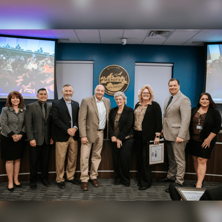 A group of eight professionally dressed people stand in front of a City of Menifee logo, smiling at the camera, showcasing the Menifee Business Spotlight for LCL Realty.