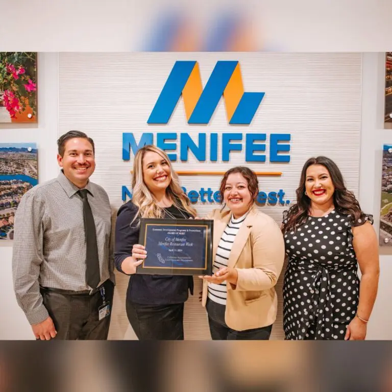 Four people are standing together in front of a "Menifee" sign, with one person holding a framed certificate celebrating Menifee's Restaurant Week.