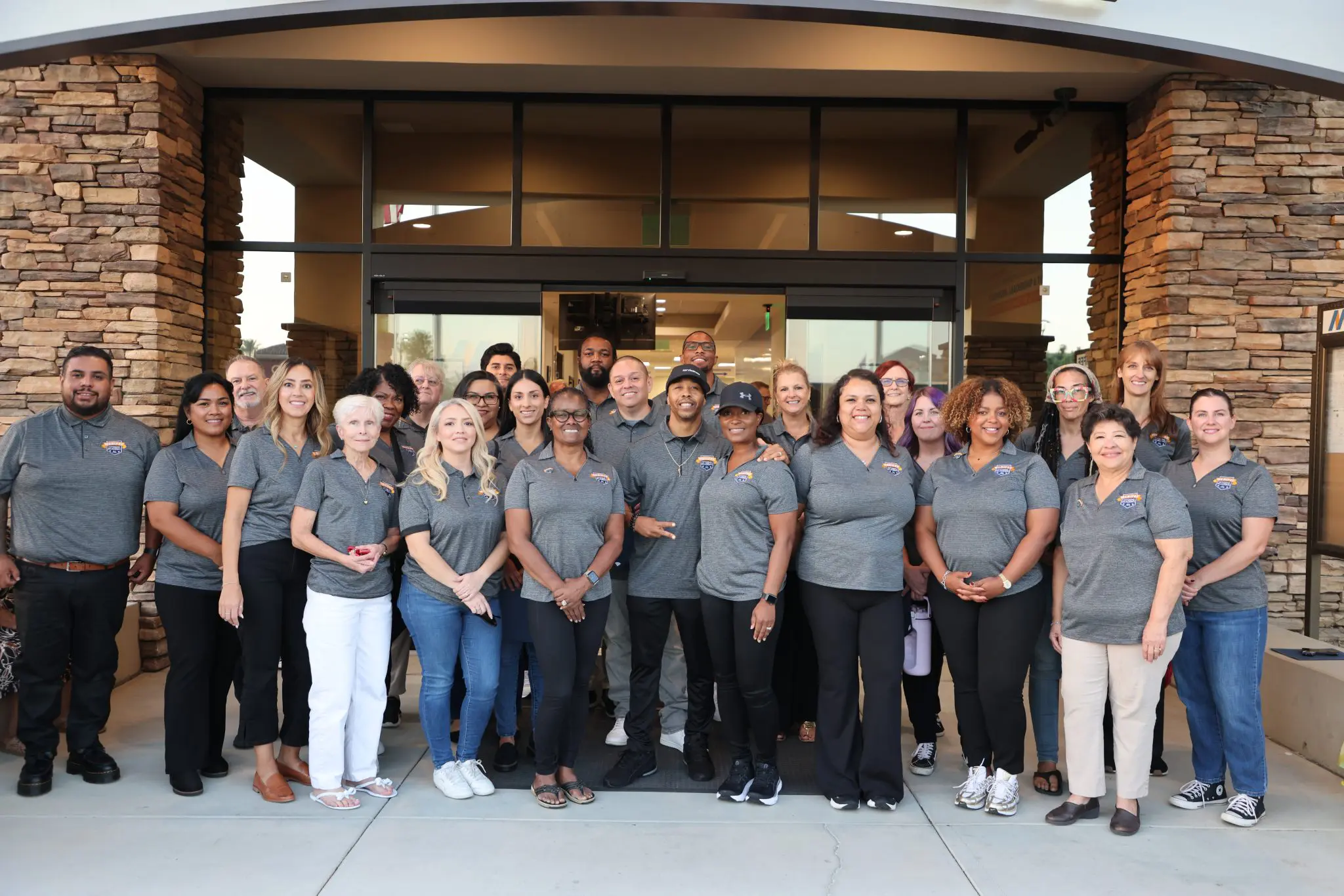 A group of adults stand in front of a building entrance, all wearing matching gray shirts and smiling at the camera, proudly representing the Menifee Citizens' Academy, a vibrant Menifee community program.